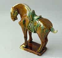 Side view of ceramic horse figurine showing sancai glaze with brown, green, and cream colors and detailed saddle.