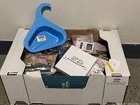 Photo showing an open box filled with assorted new items including jewelry, kitchen utensils, a blue plastic colander, greeting cards, and other household goods.