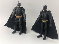 Two Batman action figures side by side in black armored suits with gold utility belts and black fabric capes.