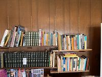 Photo showing a large assortment of books on wooden shelves including a set of green leather-bound Charles Dickens Complete Works volumes and various other titles about travel and folklore.