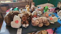 Wide shot of multiple assorted stuffed animals displayed on a table including brown and white bears, and plush ducks with tags intact.