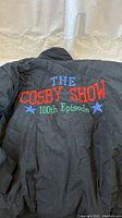 Back of black jacket with 'THE COSBY SHOW 100th Episode' embroidery in blue, red, and green thread with blue stars.