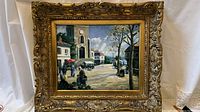 Framed oil painting showing a village street scene with people, architecture, and trees under a blue sky.