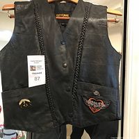 Front view of black genuine leather vest with braided leather trim, snap buttons, front pockets with Harley Davidson patches and pins.
