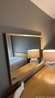 Front view of a rectangular Heywood-Wakefield mirror with light wood frame reflecting part of a bed and lamp.