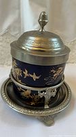 Front view showing cobalt blue porcelain decorated with gold floral and bird designs, ornate pewter lid with embossed designs and finial, set in pewter stand with ornamental feet and clip