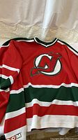 Full view of the CCM New Jersey Devils hockey jersey showing the red base color with green and white horizontal stripes and the large Devils logo patch on front.