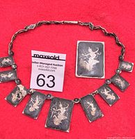 Silver Siam necklace laid out on red background showing nine rectangular engraved panels and matching brooch, highlighting detailed traditional Thai figures and patterning.