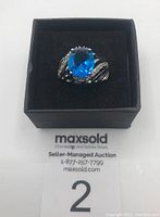 Front angle view of sterling silver ring with large blue oval stone and small clear stones, displayed on black cushion in box.