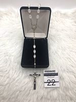 Rosary necklace with white oval beads, centerpiece medallion, and crucifix cross pendant displayed inside black jewelry box with white cushion.