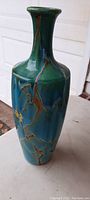 Photo showing the green and blue glazed art glass vase with brown crackle pattern and yellow decorative detail, standing upright on a surface outdoors.