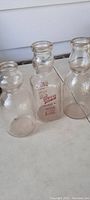 Three antique glass milk quart bottles shown side by side; one with applied color label, two with embossed lettering.