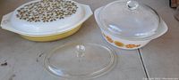 Yellow Pyrex oval casserole dish with white glass lid decorated with green leaf pattern and gold accents, alongside white round Anchor Hocking dish with orange sunflower pattern and clear glass lid