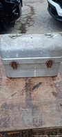 Front view of the aluminum lunch box showing two rusted metal clasps locking the box and the lunch box surface showing significant wear and some paint or rust spots.