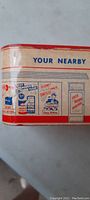 Side view of the tin coin bank showing 'Your Nearby' graphic and vintage advertising images including cans and service counters.