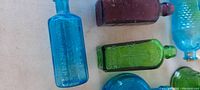 Overview of colored glass bottles showing a blue figural bottle with embossed lettering on the left, a green figural bottle in the center, and a purple bottle on top right.