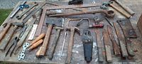 Wide view of various primitive hand tools laid out on a wooden surface including hatchets, hammers, pliers, squares, saw blades, and scraper tool showing aged and used condition with rust and wear.
