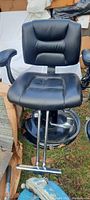 Front view of one black salon chair showing padded seat, backrest with stitched cushioning, armrests, chrome footrest and hydraulic lift mechanism on round black base.