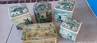 Five tin canisters grouped showing colorful Dutch scenes and graphics including windmills, boats, seagulls, and villages.