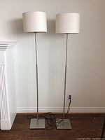 Pair of modern floor lamps with white textured drum shades and metal bases pictured against a white wall, cords visible.