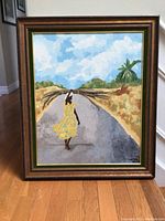 Front view of the framed painting showing woman in yellow dress walking on road with sticks on her head, framed with visible wood frame and an inner green and gold border.