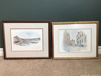 Two framed watercolor artworks hung side by side on a green wall showing boats and buildings scenes.