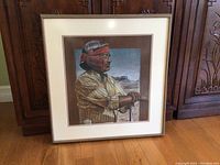 Framed art showing Native American man in profile with red headband and traditional clothing, inside a simple light frame with visible scratches.