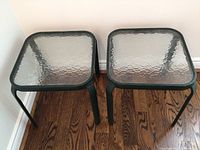 Two square patio side tables with black metal frames and textured clear glass tops placed on wood floor against white wall.