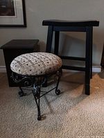 Photo displaying two stools and a dark wooden storage box placed on carpet. One stool has a round cushioned seat with patterned fabric and black metal decorative legs. The other stool is black wood with a simple flat seat. Storage box visible in background with damaged edge.