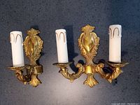 Pair of vintage brass Baroque style sconces shown mounted with candle-shaped holders featuring white sleeves with gold wax drip design.