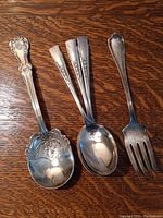 Five antique silver serving pieces laid out on wooden surface showing ornate and floral designs, sizes, and types of utensils.