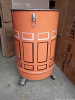 Side view of the orange steel canister showing geometric pattern and caster wheels.