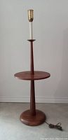 Full view of vintage Mid-Century Modern walnut floor lamp with round table built-in and tall slender lamp holder, standing on concrete floor against white wall.