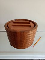 Front and side perspective of the round teak ice bucket with lid on, showing wood grain and carved handle slots on lid, along with pencil for scale