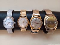 Front view of four vintage men's watches on black watch holder showing dial faces and metal bands