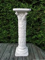 Full view of white painted plaster Greco-Roman style column plant stand outdoors on wooden deck in front of greenery.