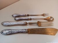Full set of four silver handle serving utensils laid out on white surface including knife, fork, spoon, and small two-pronged fork.