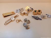 Top view of collection displaying multiple pairs of cuff links, a gold-tone tie bar with white center, and a ribbed money clip, all laid out on a plain surface.
