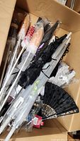 Box containing multiple parasols with some still in plastic packaging, visible black lace parasols, and lace fans.