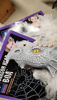 Silver dragon head with yellow eye attached to white feather boa, in original packaging visible.