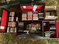 Photo of a black and red carrying case holding multiple 8 track tapes and cassette tapes inside compartments, showing variety of tapes and labels.