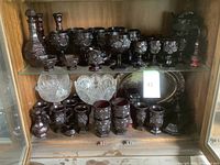 Wide view of multiple dark red Avon Cape Cod glassware pieces arranged on shelves, including goblets, decanters, small bowls, covered butter dish, and clear cut glass bowls and plates.