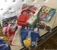 Photo showing multiple XL T-shirts with Looney Tunes designs, NFL Super Bowl XIII T-shirt, and Avon collectible beer steins in gift boxes along with Avon Christmas potpourri box on a mattress.
