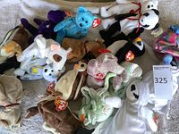 Overview of assorted Ty Beanie Babies plush toys laid out on a textured light surface, showing multiple colorful characters with attached red Ty heart tags.