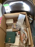 Brown metal folding chair behind a box containing assorted vintage matchbooks and small collectibles including lighters, pins, and paper ephemera.