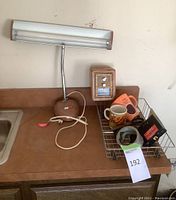 View of brown desk lamp, small wooden combination safe, two mugs, metal wire basket holding scissors and miscellaneous items