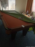 View of the pool table with green cover partially pulled back, showing orange-toned red felt and wooden frame.