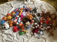 Group of various small stuffed animals laid out on a white sheet, showing a mix of character and animal plush toys including multiple GUND and Beanie Babies items.