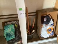 Two cardboard boxes with folded T-shirts, one baseball cap, and a plastic bucket holding wooden-handled hand tools.