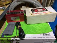 Photo showing the isolator unit, remote control device, and original box on a green cloth with surrounding objects.